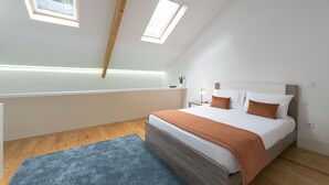 Een slaapkamer met een schuine kap, dakramen, een bed met houten hoofdbord, een nachtkastje met lamp en een blauw vloerkleed.