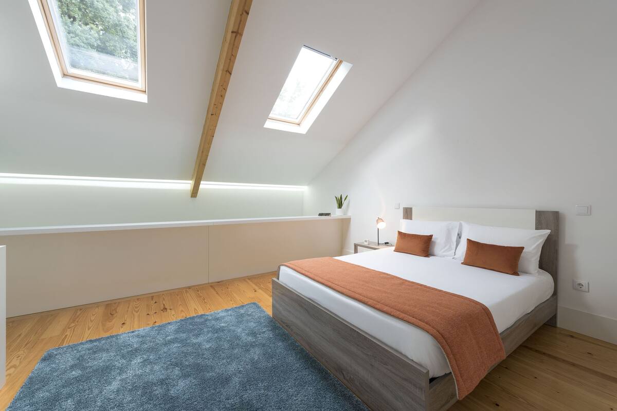 Een slaapkamer met een schuine kap, dakramen, een bed met houten hoofdbord, een nachtkastje met lamp en een blauw vloerkleed.