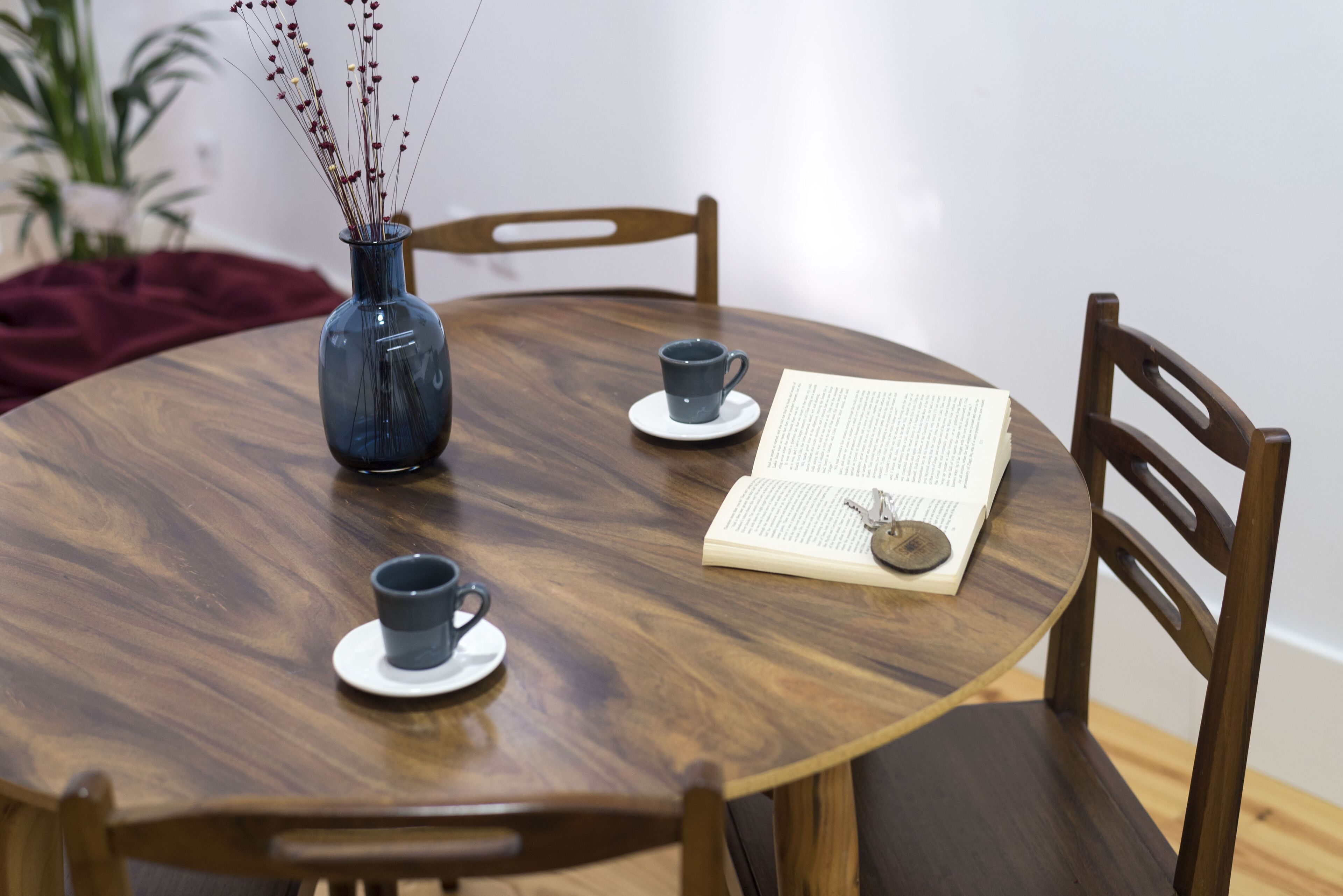 Una mesa redonda de madera con un florero azul, dos tazas y un libro abierto.