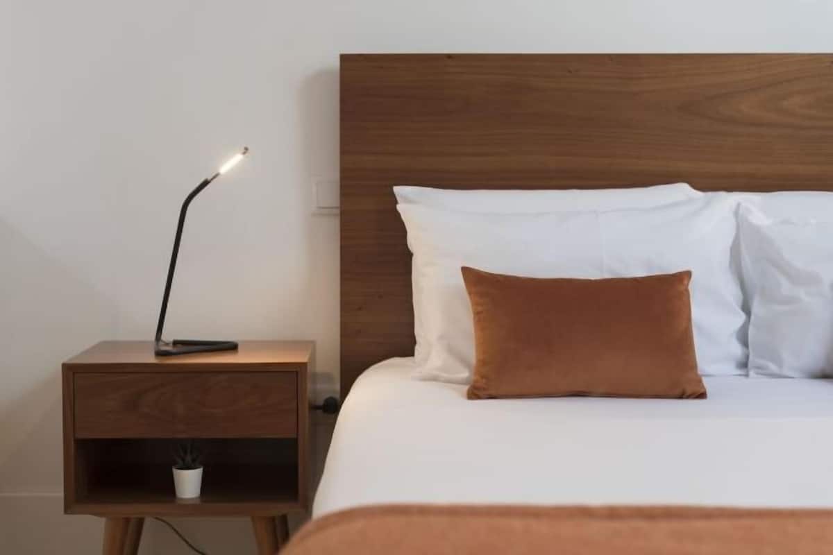 Een net opgemaakt bed met witte en bruine kussens, een bedlampje en een klein plankje met een plant.