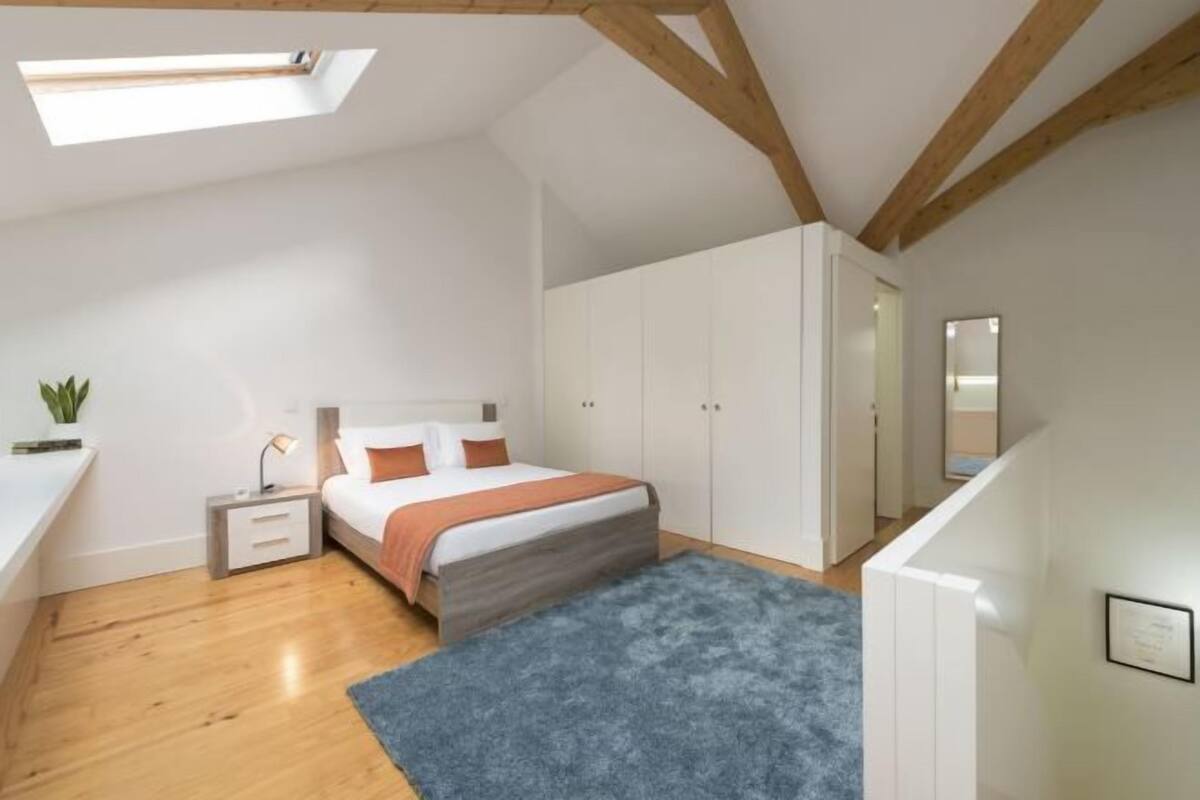 Een slaapkamer met dakraam, een bed met blauw vloerkleed, houten balken en witte kasten.