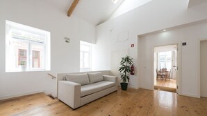 Een ruime kamer met een houten vloer, een witte bank, een kamerplant en uitzicht op een eetgedeelte door een geopende deur.