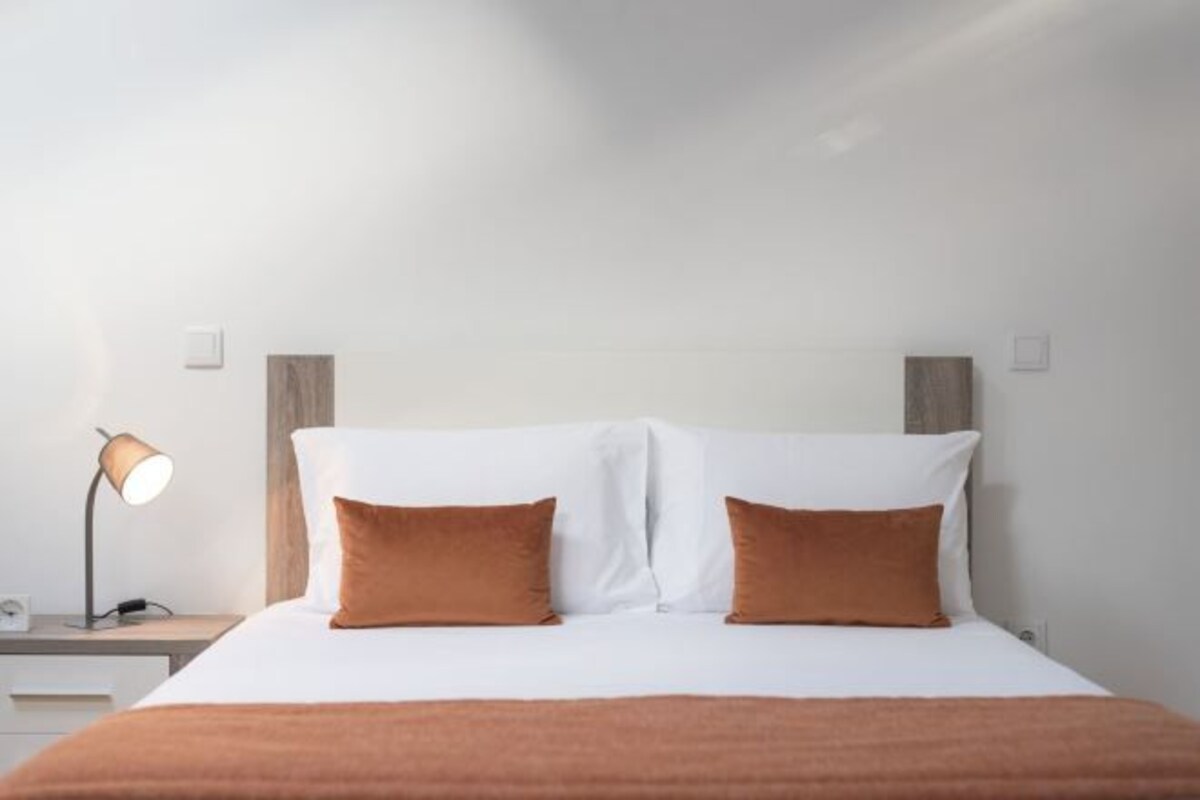 A hotel room with a bed, a nightstand, a lamp, and a wall-mounted light switch.