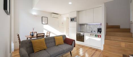 Un apartamento moderno con sofá gris, suelo de madera, cocina con armarios blancos y comedor con mesa y sillas.