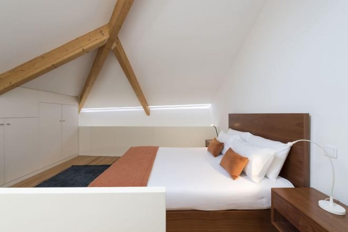 Ein Schlafzimmer mit schrägen Wänden, einem Bett mit weißer Bettwäsche, einem Holz-Kopfteil, einem Nachttisch und einer Stehlampe.