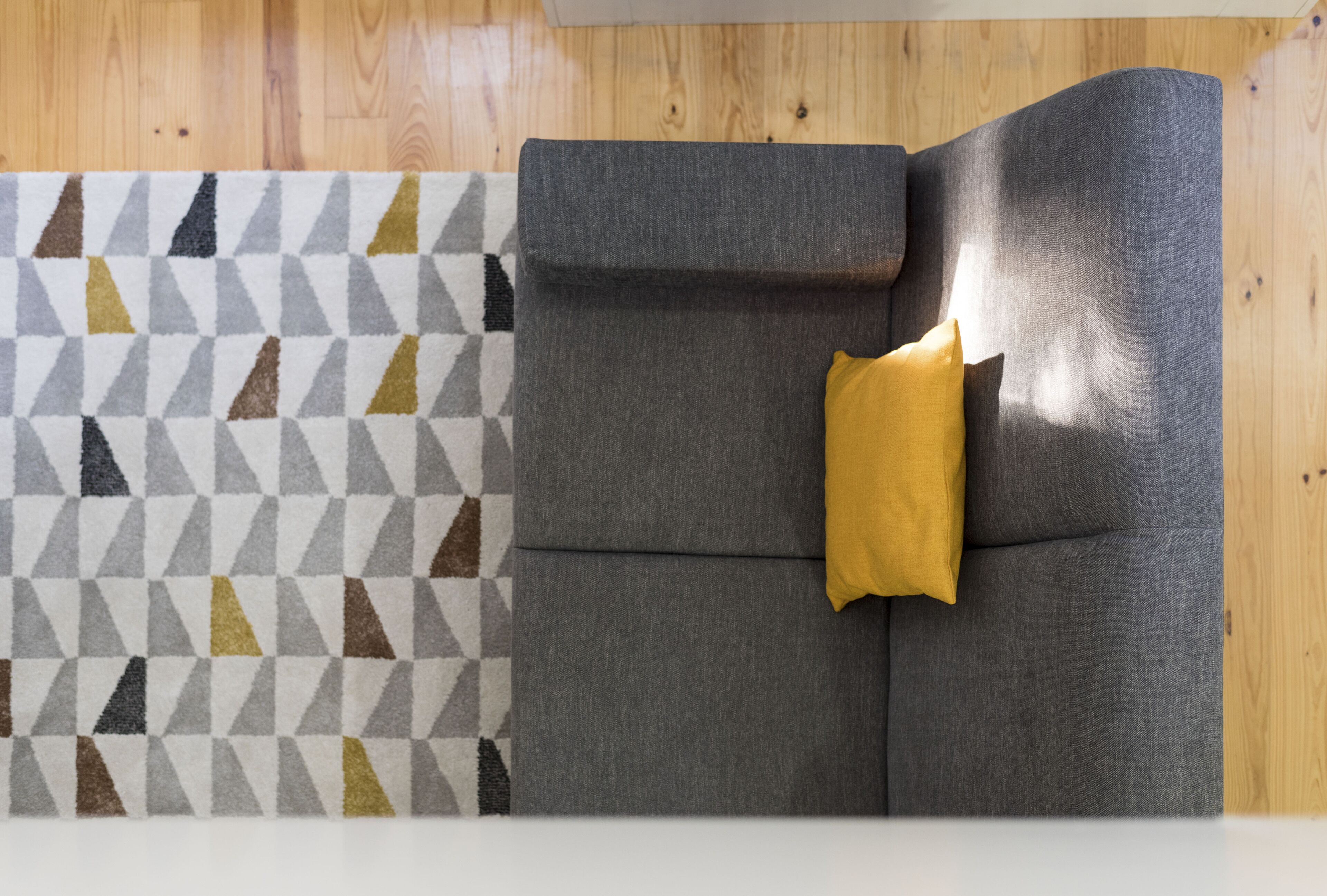 A grey sofa with a yellow pillow against a wooden floor, next to a patterned rug.