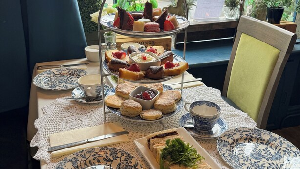 Uma mesa posta com diversos sanduíches, scones e doces, com vista para uma rua da cidade através de uma janela.