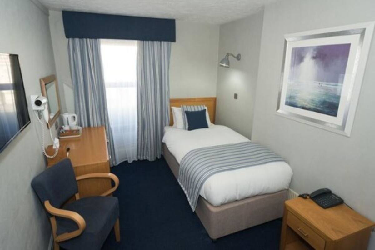 A hotel room with a bed, a desk with a chair, a bedside table with a lamp, a window with curtains, and a framed picture on the wall.