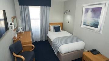 A hotel room with a bed, a desk with a chair, a bedside table with a lamp, a window with curtains, and a framed picture on the wall.