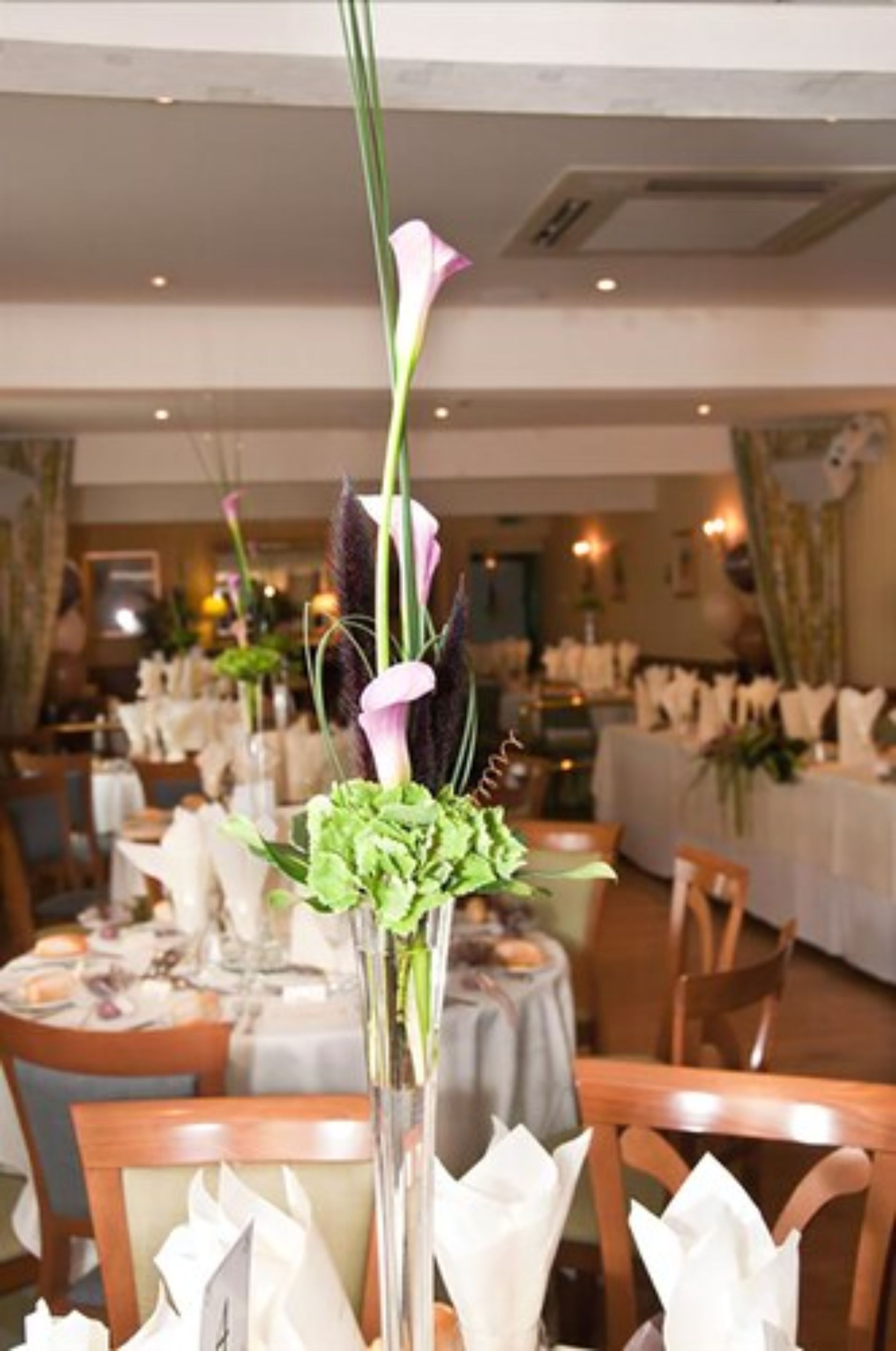 A dining area with tables set for service, featuring a centerpiece with flowers and a tall vase.