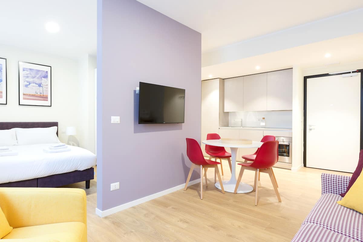 A modern hotel room with a bed, a TV, a dining area with red chairs, and a kitchen.