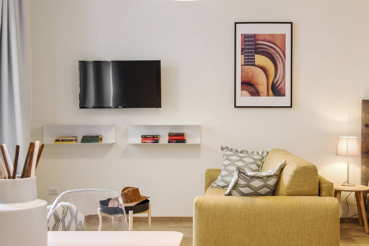 A hotel room with a flat-screen TV, a framed picture, a bookshelf, a sofa, a bed, and a small table with a lamp.