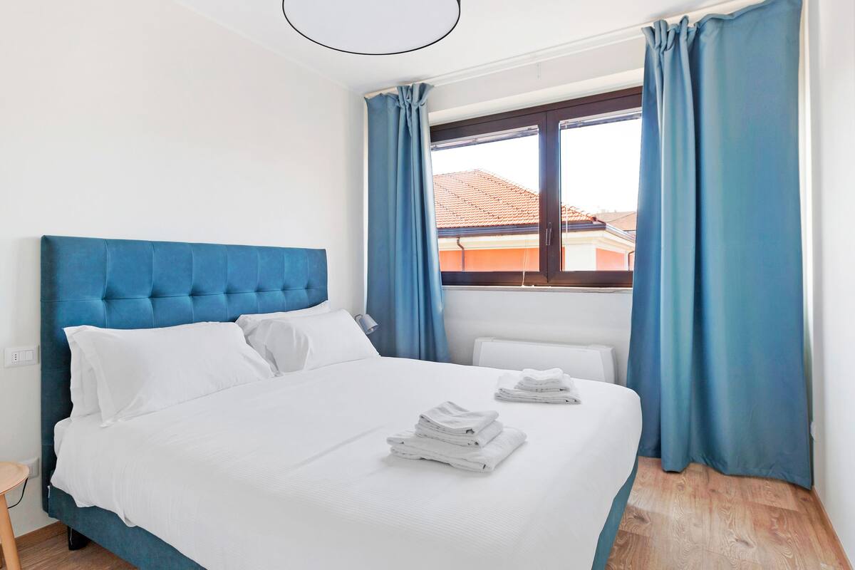 A neatly made bed with white linens and a blue headboard, a small bedside table with a lamp, and a window with blue curtains.