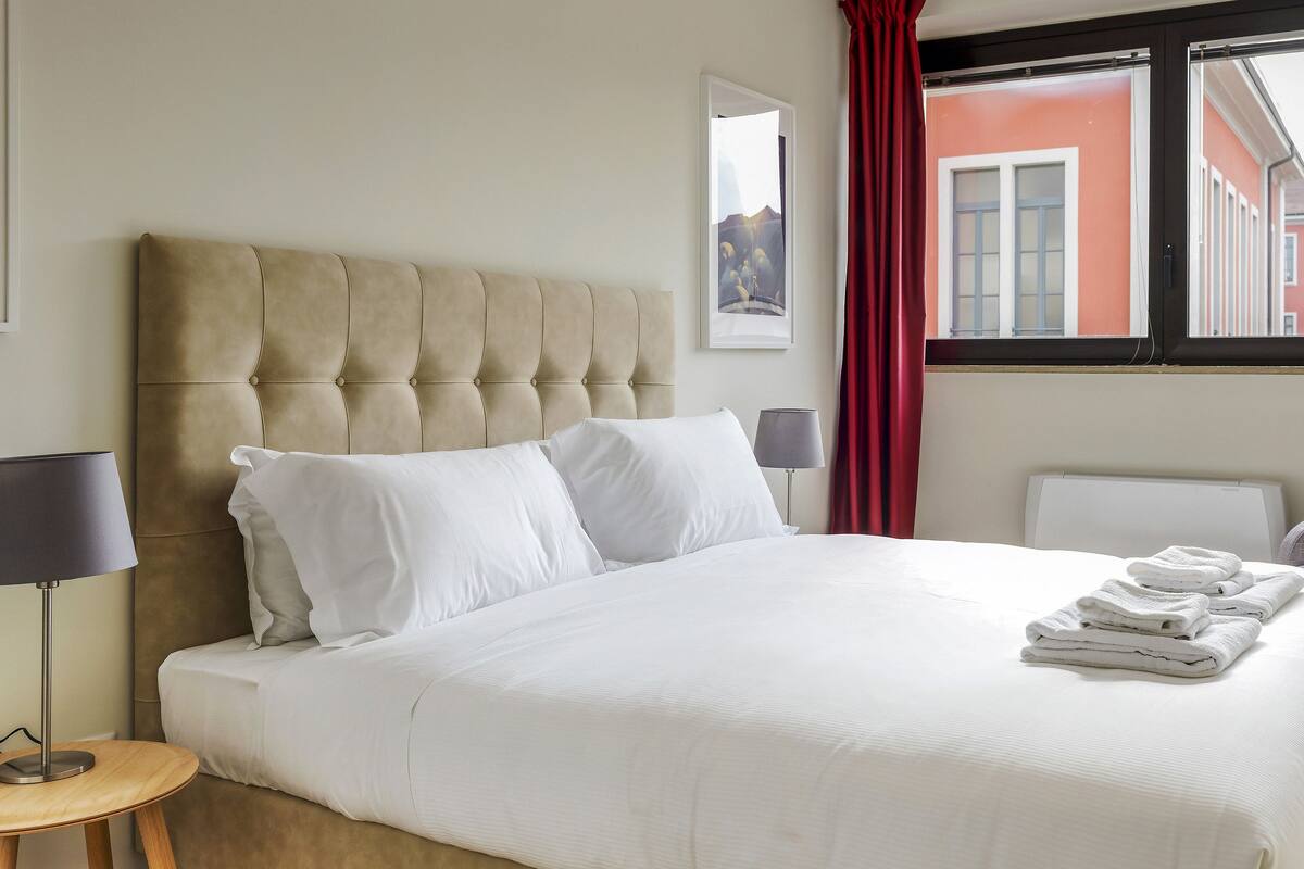 A neatly made bed with white linens, a tufted headboard, and a window with red curtains.