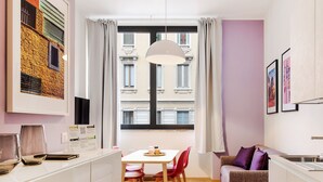 A modern living room with a dining area, featuring a large window with curtains, a hanging light fixture, and a view of buildings outside. - Italianway   - Bergonzoli (Milan)