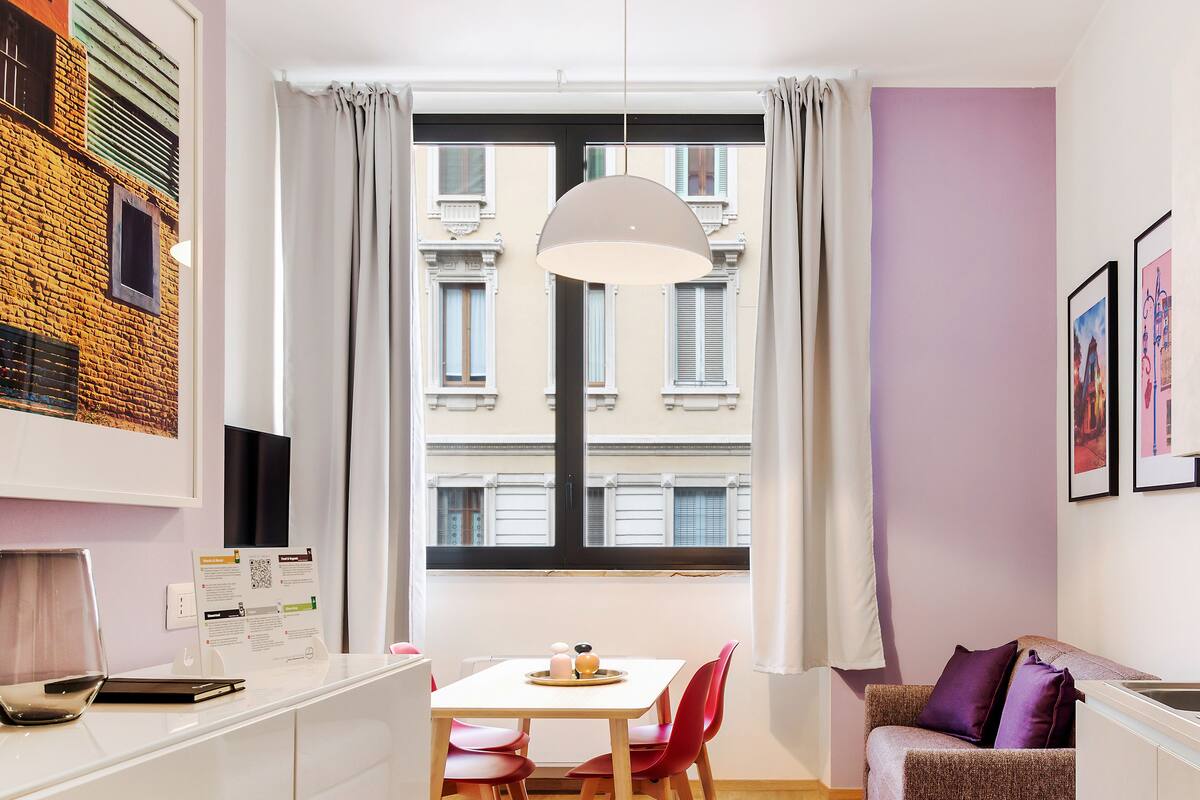 A modern living room with a dining area, featuring a large window with curtains, a hanging light fixture, and a view of buildings outside.