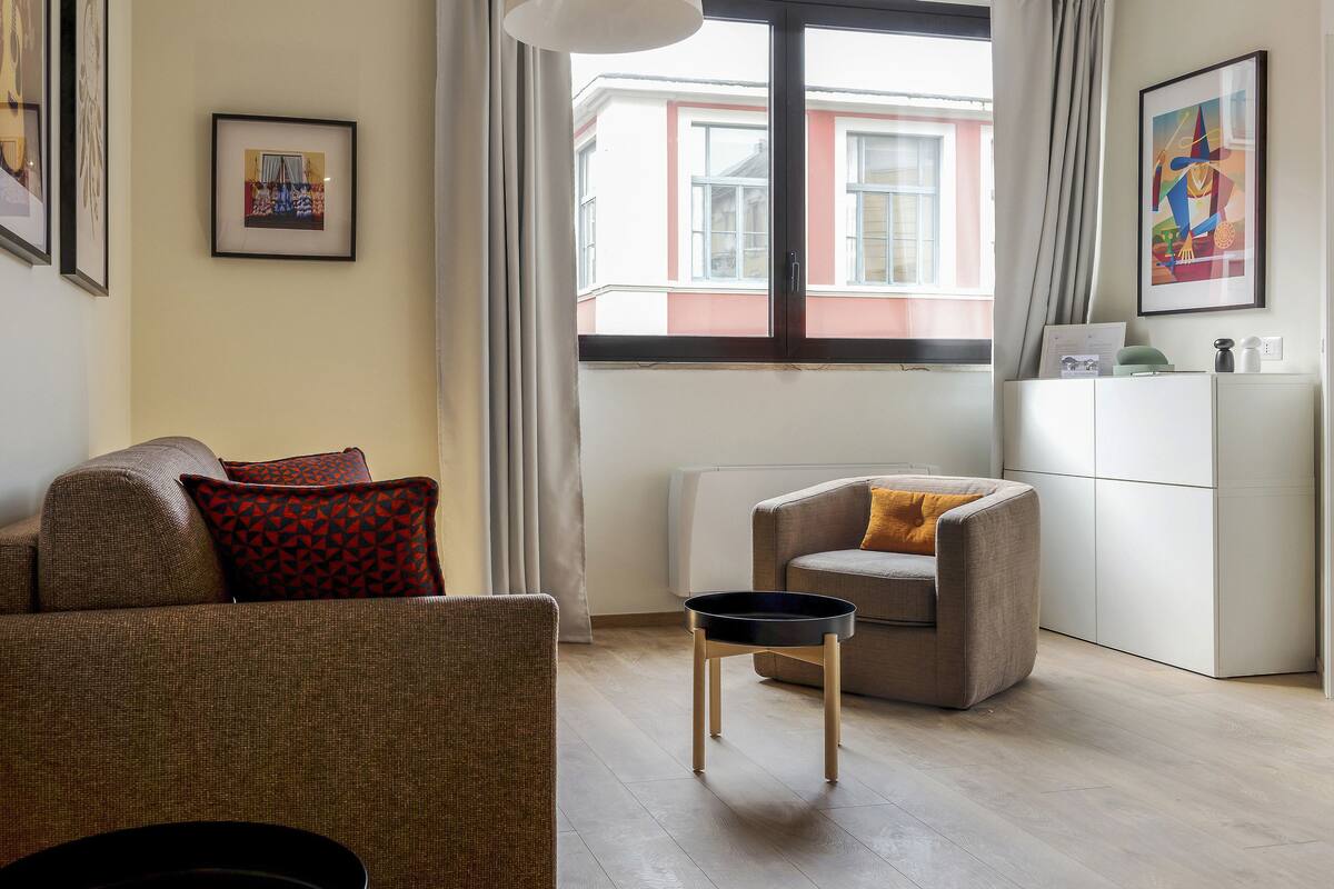 A living room with a sofa, armchair, and a small table. There are framed pictures on the wall and a window with a view of buildings outside.