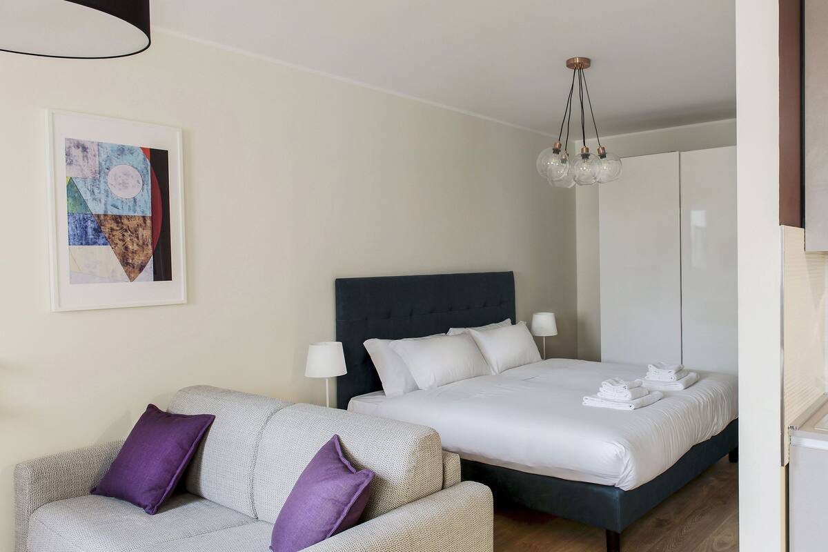 A hotel room with a bed, a sofa, a painting, and a kitchenette.