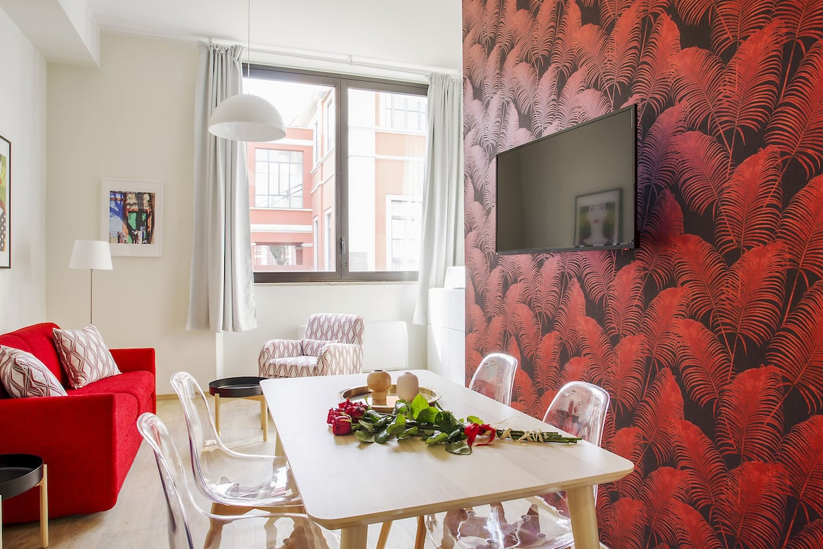 A modern living room with a red sofa, a white dining table with chairs, a flat-screen TV mounted on the wall, and a window with curtains.