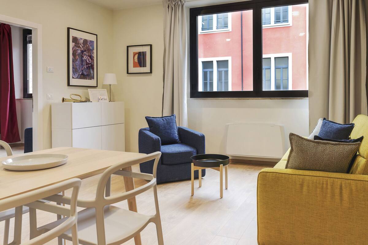 A modern living room with a dining area, featuring a sofa, chairs, a table, and artwork on the walls.