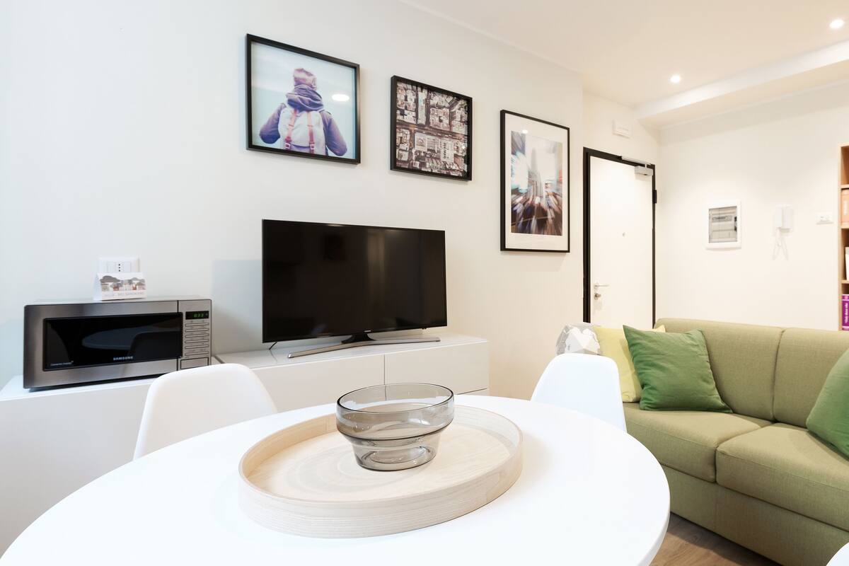 A modern living room with a flat-screen TV, a dining table with a glass centerpiece, a green sofa, and a bookshelf with books.