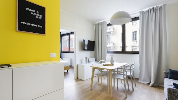 A modern hotel room with a yellow accent wall, a dining area with a table and chairs, a bed, a TV mounted on the wall, and a window with curtains. - Italianway   - Bergonzoli (Milan)