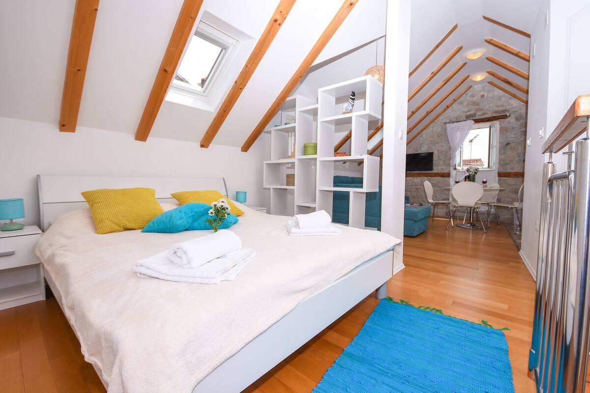 A bedroom with a bed, white walls, wooden beams, a skylight, a bathroom with a stone wall, and a dining area.