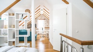 A modern loft bedroom with a wooden ceiling, white built-in shelving, a bed with a rolled-up white blanket, and a dining area with a stone wall and white furniture. - Tragos Lemon Tree (Trogir)