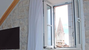 A room with a stone wall, a wooden ceiling beam, a window with a view of a building, a table with a glass bowl of lemons, a bottle, and a mug. - Tragos Lemon Tree (Trogir)