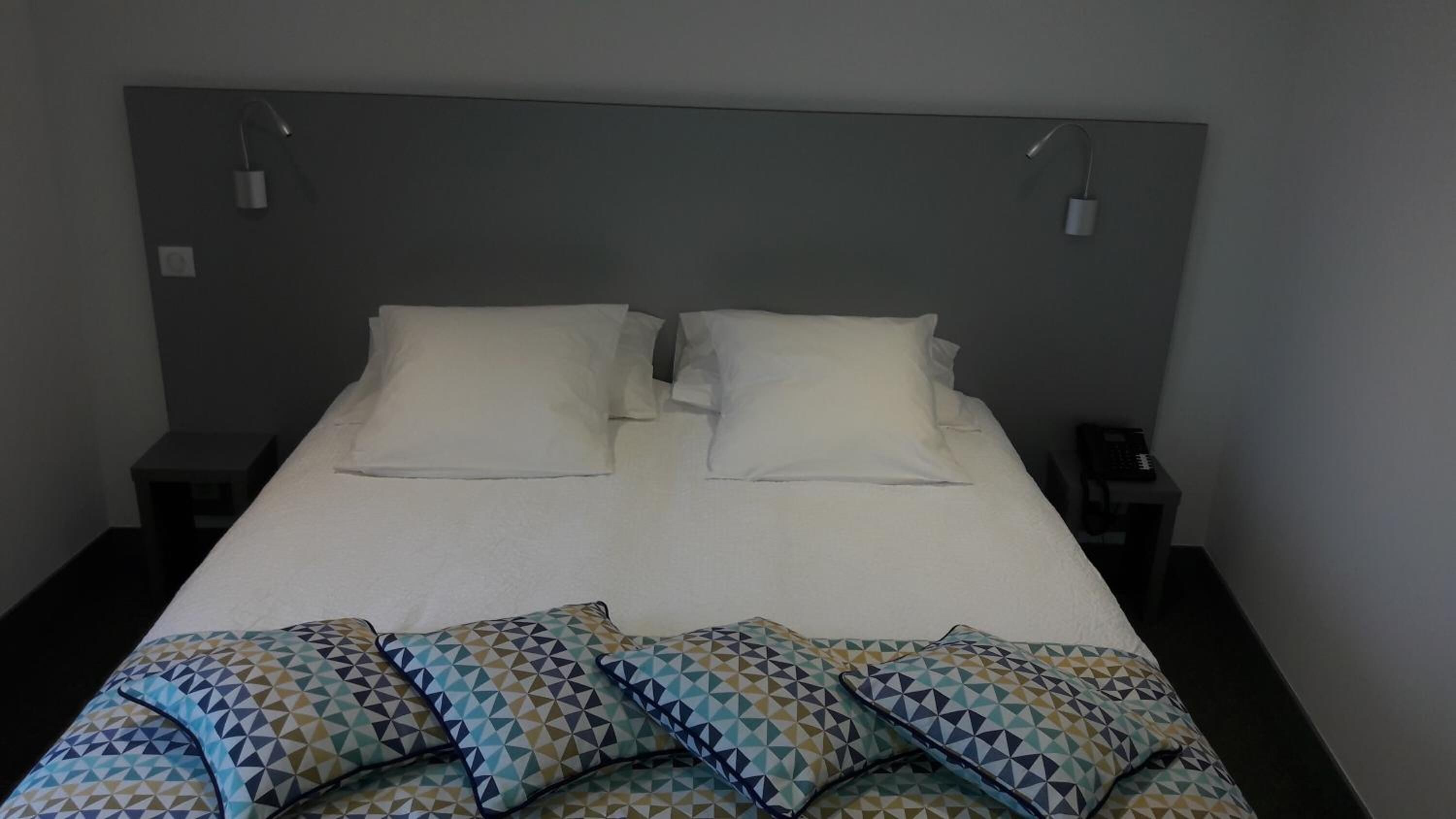 Un lit bien fait, avec du linge de lit blanc et des coussins aux motifs géométriques, deux tables de chevet avec des lampes et un mur sobre.