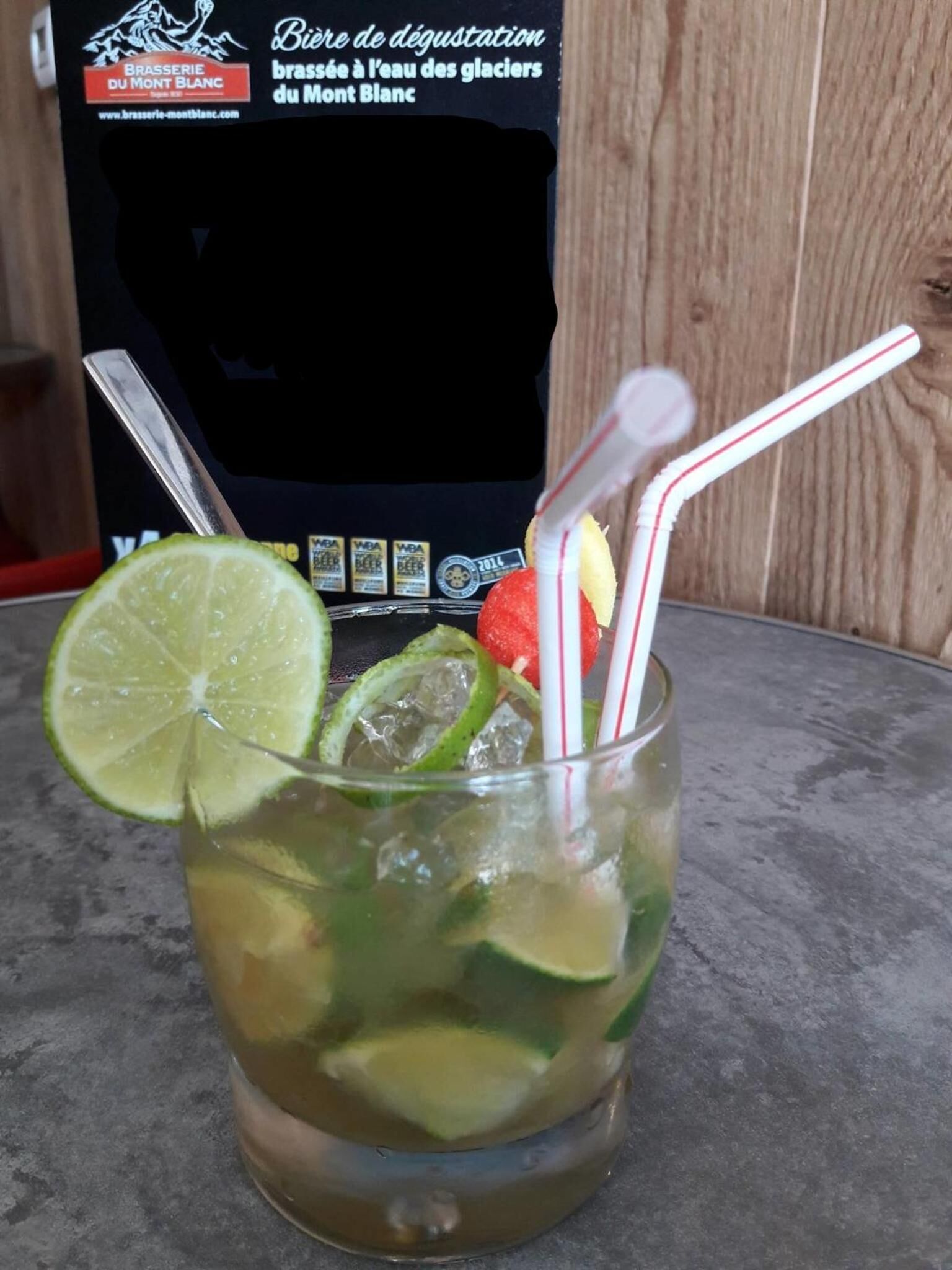 A glass of a beverage with lime and mint, with a straw and a slice of watermelon, on a table with a wooden background.