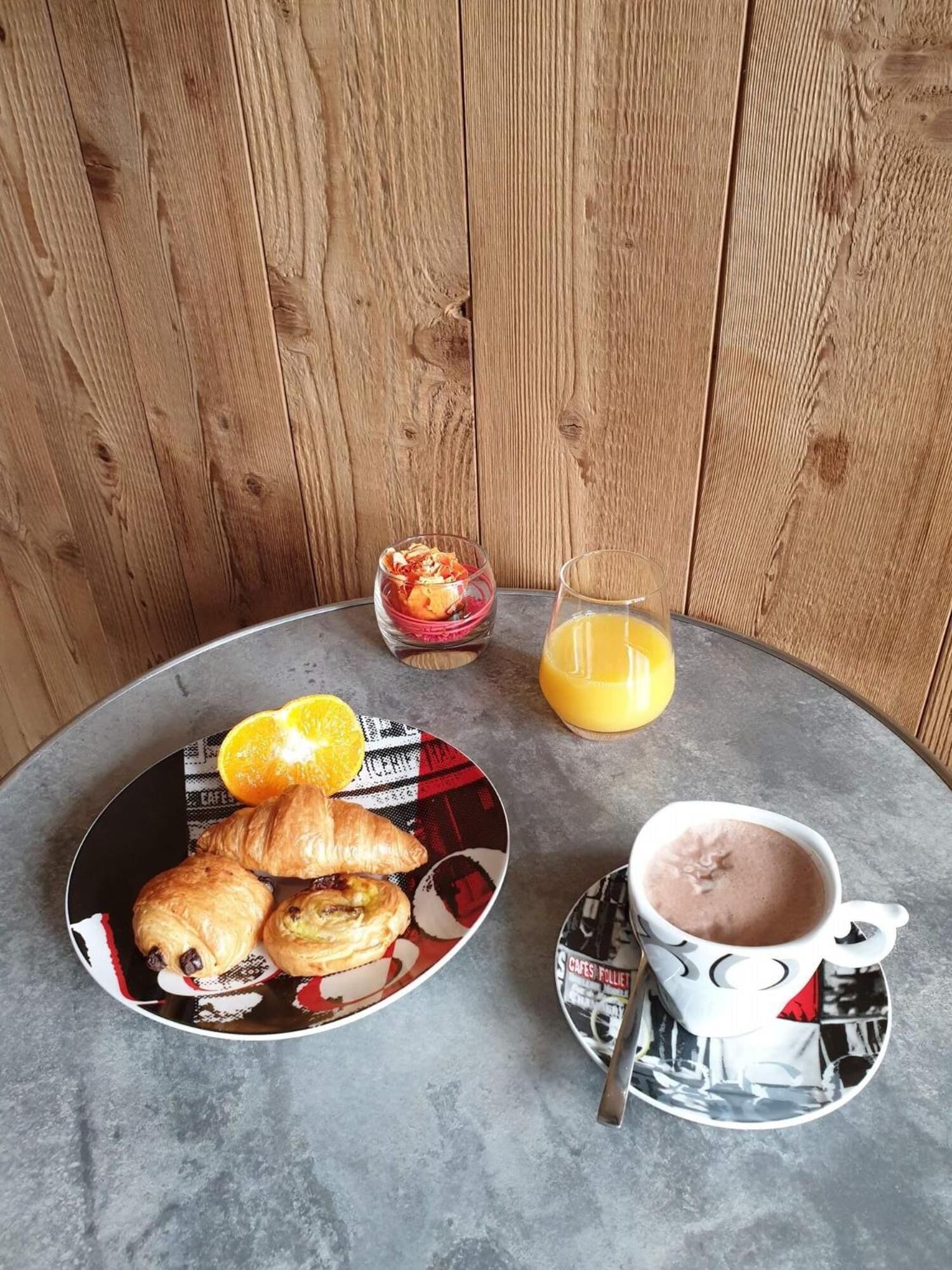 Un desayuno con croissants, un bol de fruta, un vaso de zumo de naranja y una taza de chocolate caliente sobre una mesa redonda.