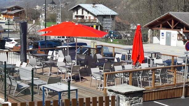 Un espace extérieur aménagé avec des tables et des chaises, un parasol rouge, et une vue sur les bâtiments et les montagnes environnants.