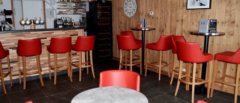 Un coin bar avec des tabourets rouges, une table ronde et un mur en bois orné d’une horloge et d’un tableau encadré.