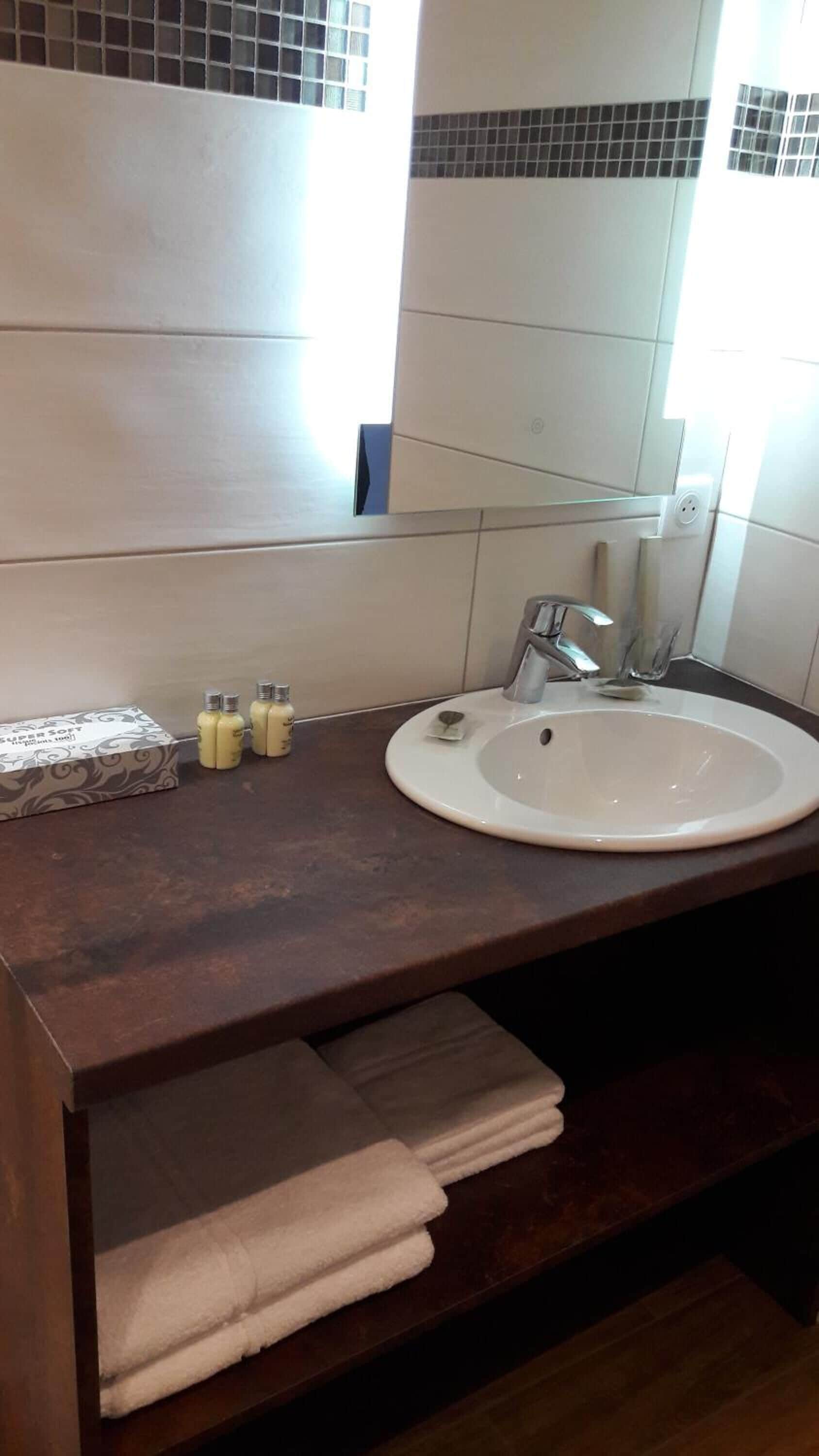 A bathroom with a white sink, dark brown countertop, and a mirror above it. There are three bottles of lotion on the counter and a stack of white towels below the mirror.