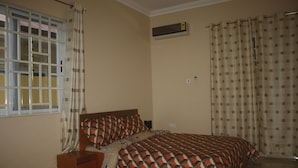 Un dormitorio con cama, mesita de noche, cómoda, ventana con cortinas y un equipo de aire acondicionado.