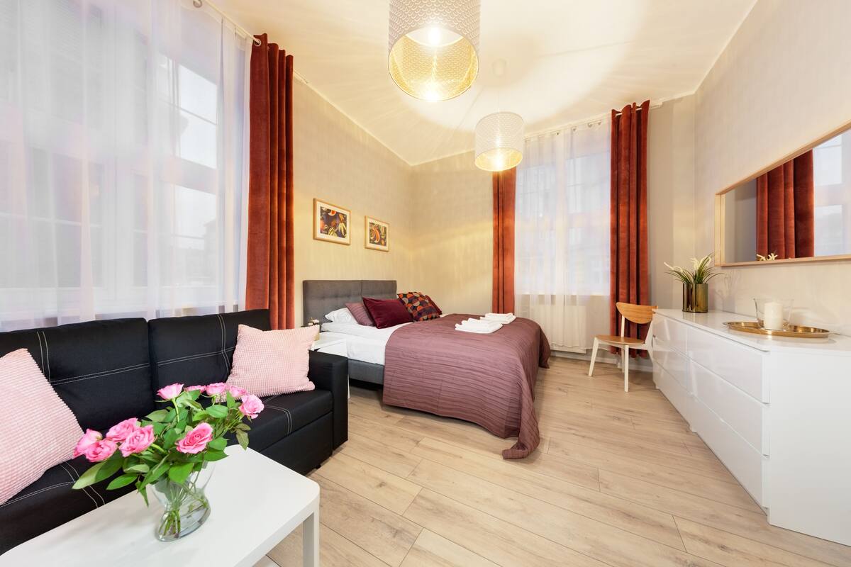 A modern hotel room with a bed, a sofa, a dining area, and a large window.