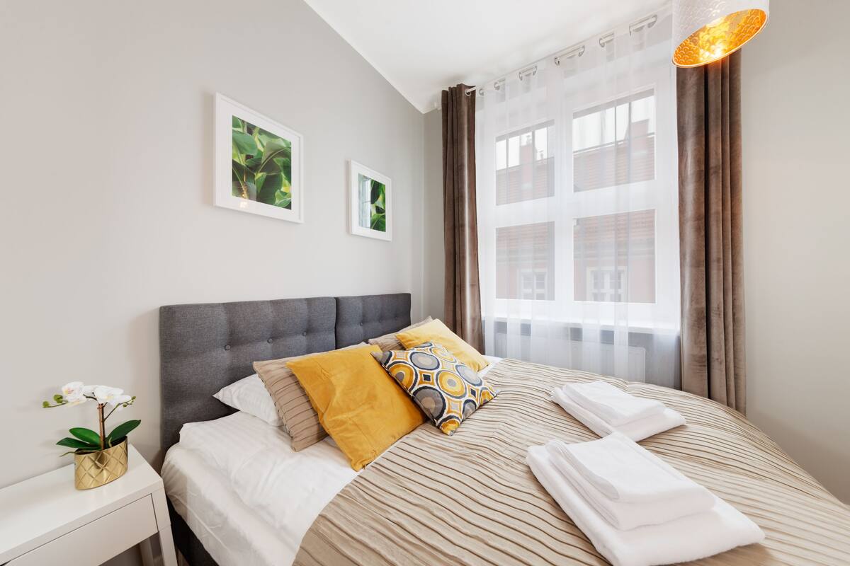 A neatly made bed with a striped bedspread, a grey upholstered headboard, and two pillows. There is a small bedside table with a vase of flowers and a framed picture on the wall.