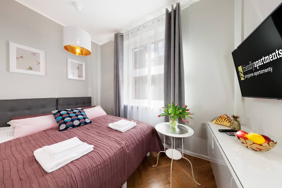 A modern hotel room with a neatly made bed, a small table with a vase of flowers, and a fruit basket on a white dresser.