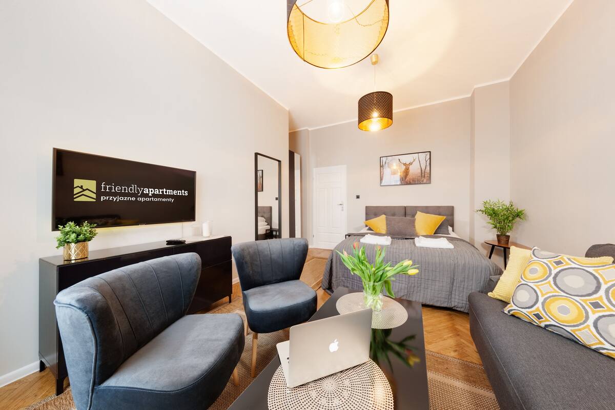 A modern living room with a sofa, two armchairs, a coffee table with a laptop, a TV, and a bed with a headboard.