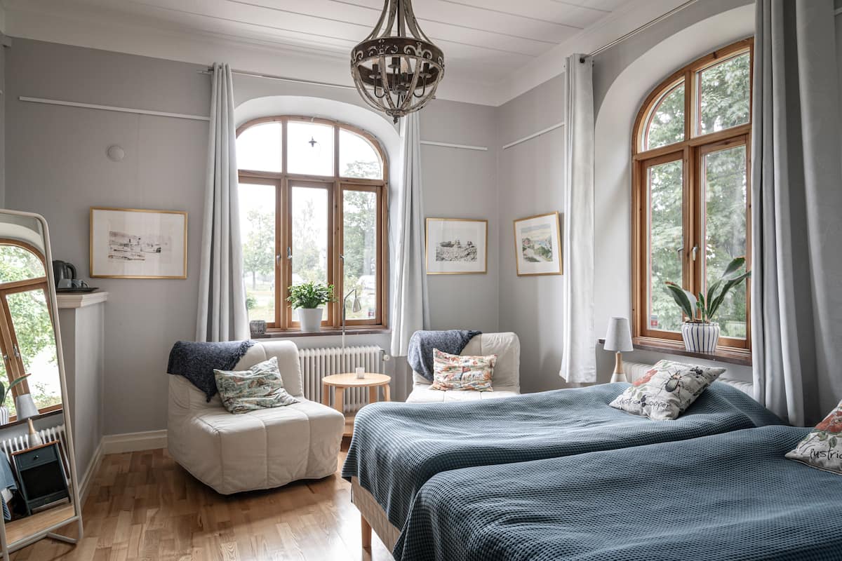 Ein Schlafzimmer mit einem großen Bett, zwei Sesseln, einem kleinen Tisch und einem Heizkörper. Es gibt große Fenster mit Vorhängen, eine Pendelleuchte und gerahmte Bilder an den Wänden.