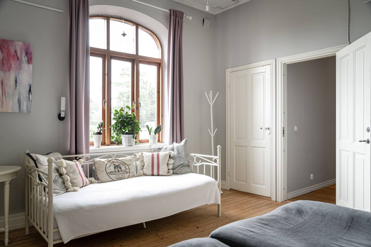 Ein gemütliches Zimmer mit einem weißen Bett, einem Fenster mit Vorhängen, einer Topfpflanze und einer weißen Tür.