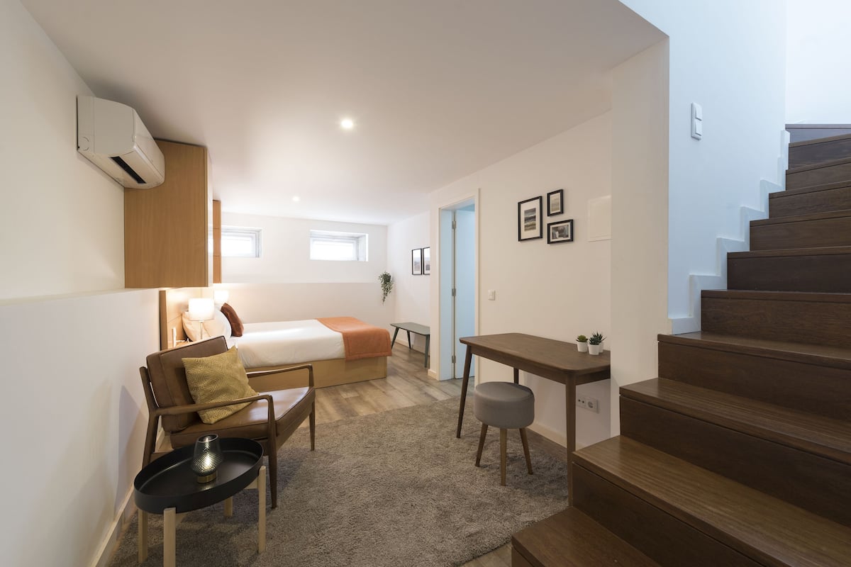 Ett kompakt hotellrum med en säng, ett skrivbord, en stol, ett litet bord och en trappa.