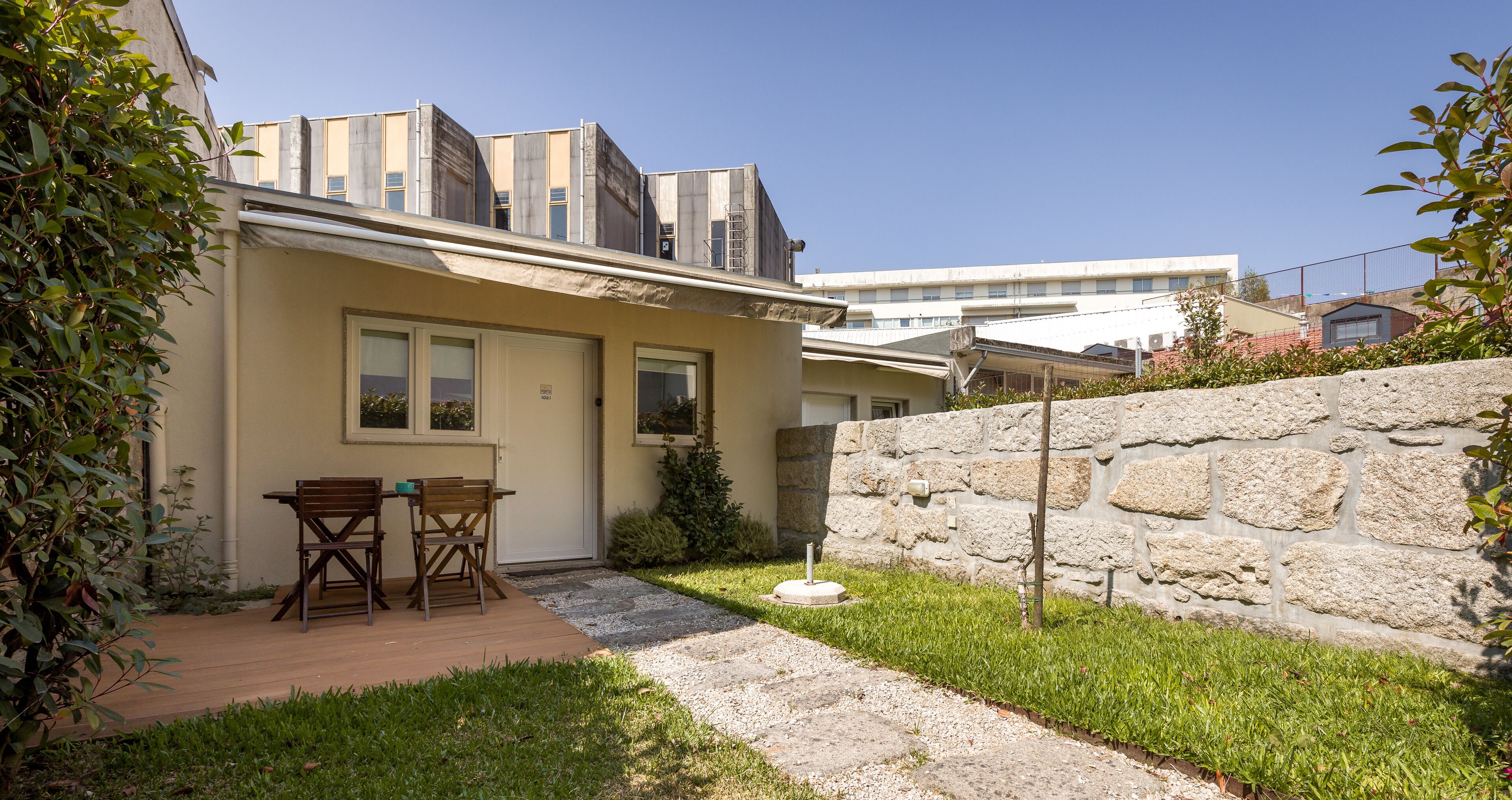 Un edificio residencial con un muro de piedra, una terraza de madera con sillas y un pequeño jardín.