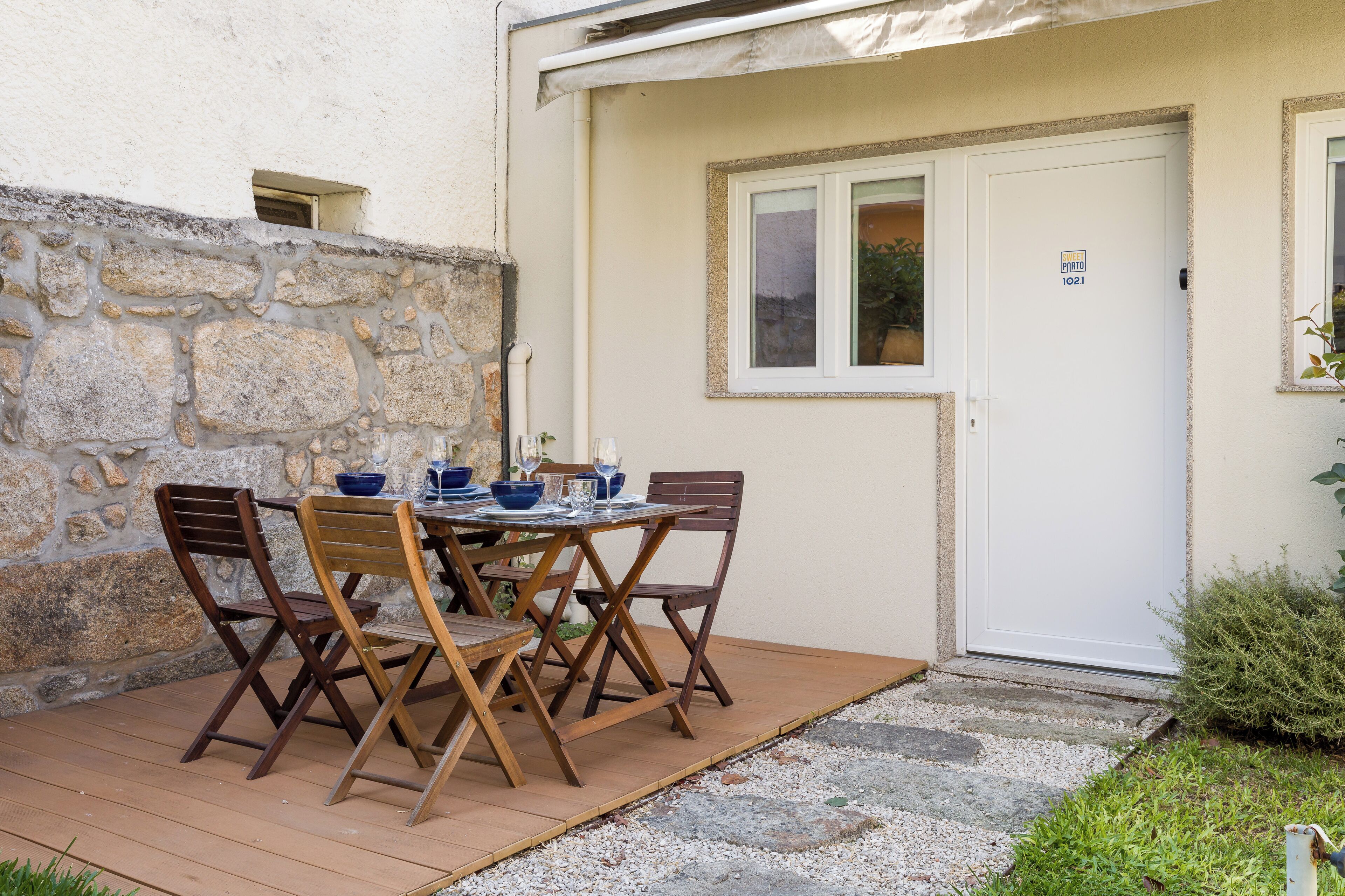 A small outdoor dining area with a table and chairs, a stone wall, and a door with a window.