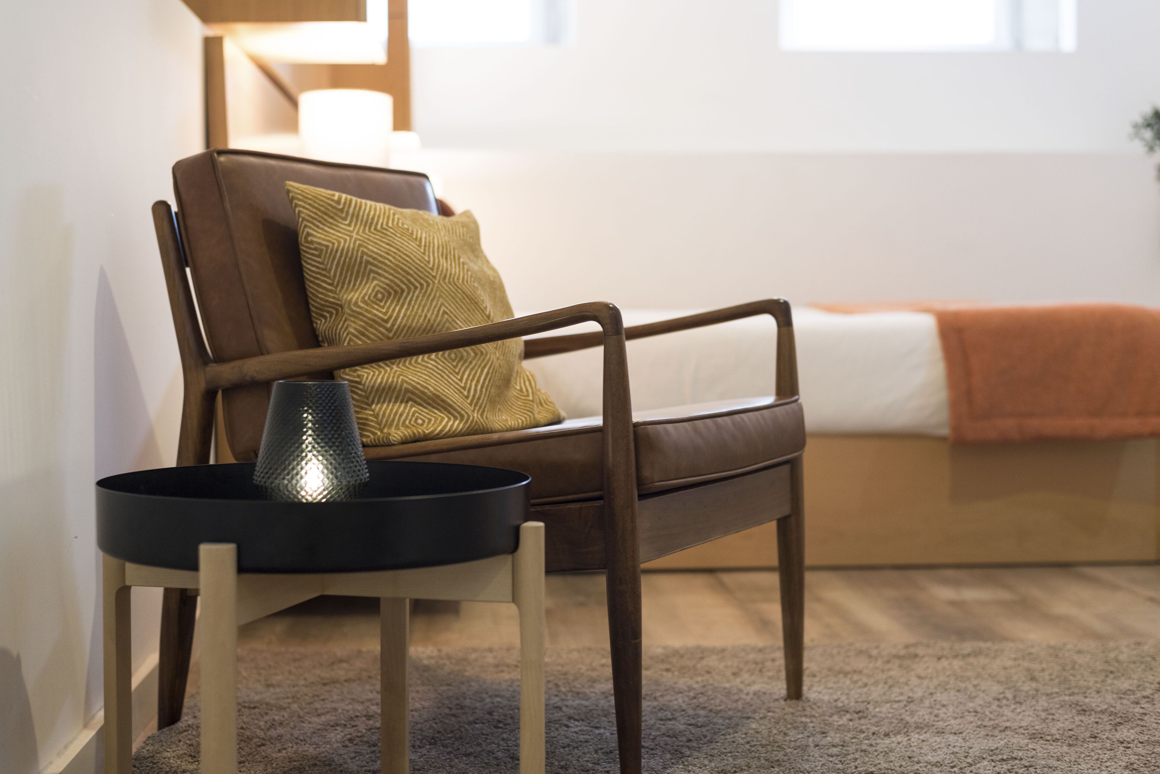 A modern living room with a brown leather sofa, a small round table with a lamp, and a window with a view of the outdoors.