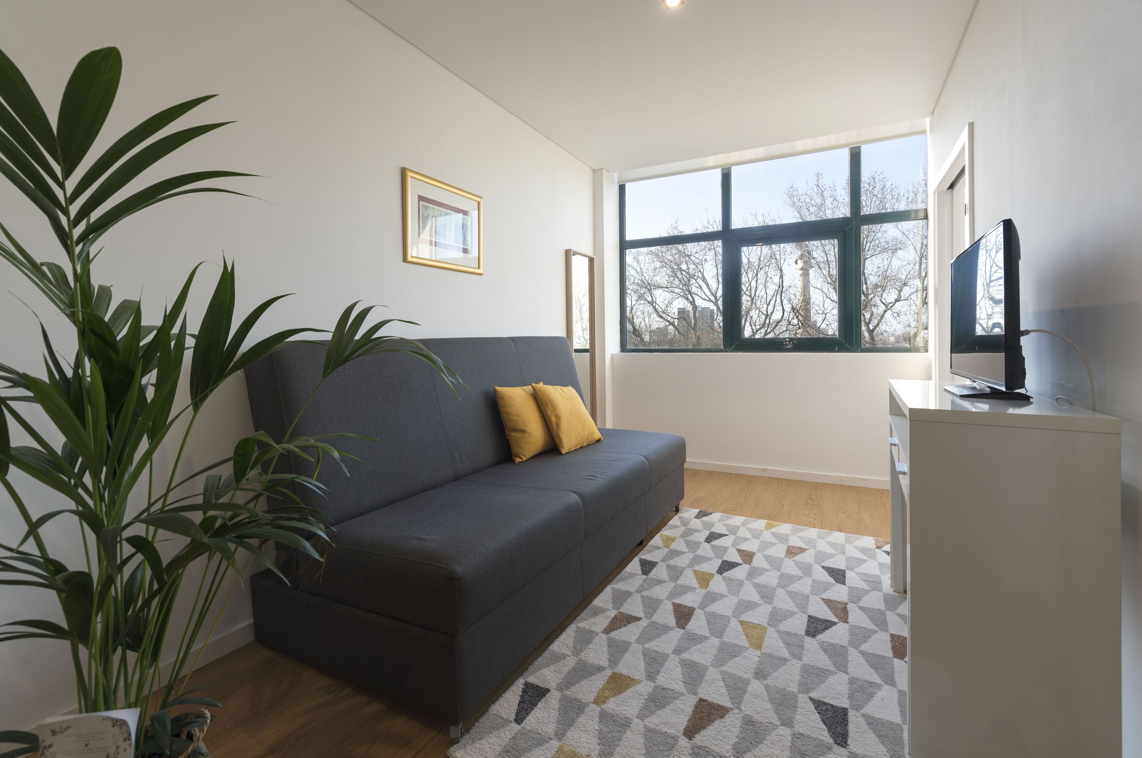 Ein modernes Wohnzimmer mit einem dunkelgrauen Sofa, einem gemusterten Teppich und einem großen Fenster mit Blick auf Bäume und Gebäude.