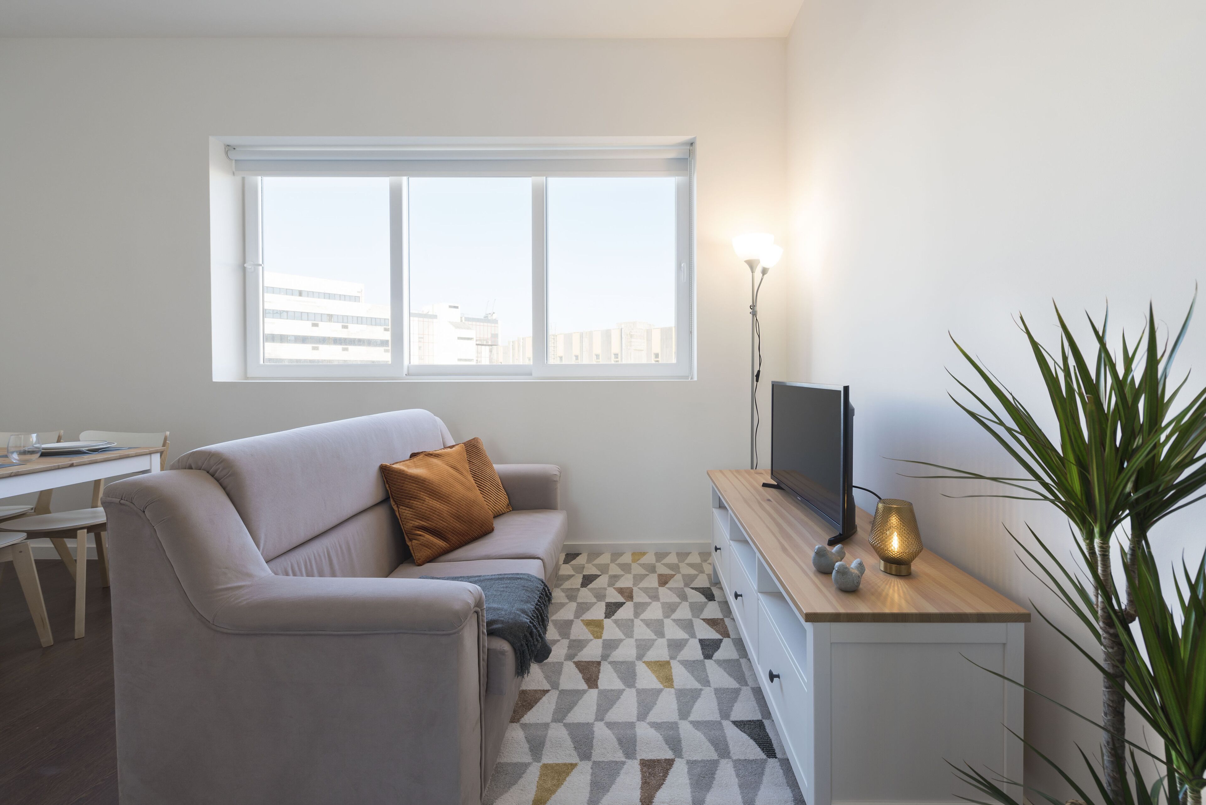 Een moderne woonkamer met een bank, een televisie, een lamp en uitzicht op de stad door een groot raam.
