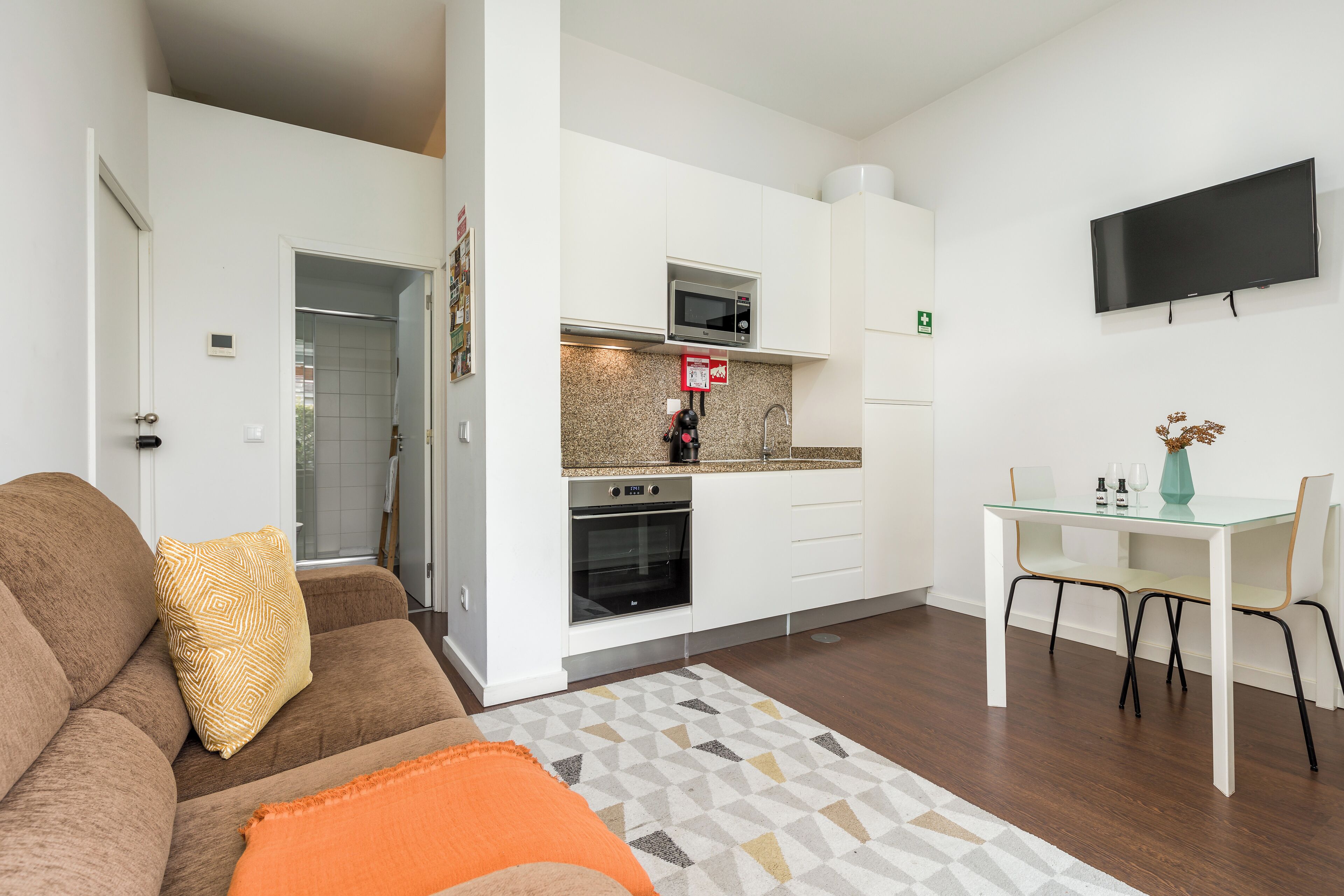 Un espacio habitable compacto con kitchenette, un sofá con cojines, una mesa de comedor con sillas y un televisor de pantalla plana montado en la pared.