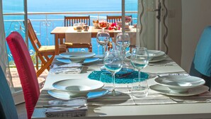 Una mesa de comedor preparada para cuatro personas, con platos blancos, copas de vino y un mantel individual azul. La mesa está ubicada cerca de una ventana con vistas al mar.